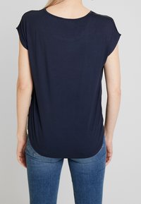 Blond person iført en navyblå kortærmet bluse med rundet kant, parret med lysvaskede blå jeans, vist fra ryggen.