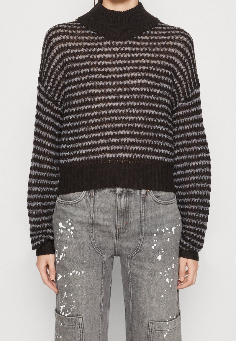 Maglione a strisce in bianco e nero con collo alto, maniche ampie e una vestibilità corta, abbinato a pantaloni grigi con macchie di vernice.