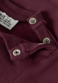 Maglietta per neonato bordeaux con colletto a coste, chiusura a pressione e texture morbida. Etichetta di cura visibile sul lato sinistro.