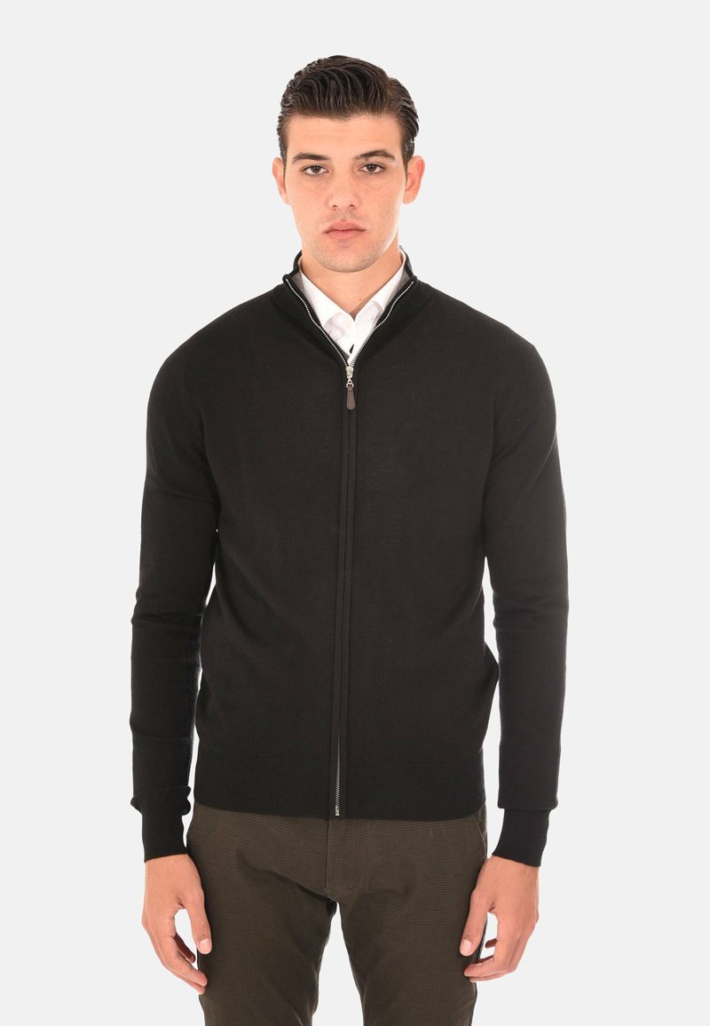 Maglione zip nero con colletto alto, realizzato in tessuto morbido, con un design aderente e polsini e bordo a coste. Indossato sopra una camicia bianca.