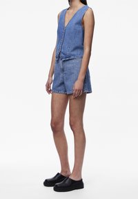 Vrouw die een mouwloos spijkervest met knopen en gestikte spijkershorts draagt, met zwarte platform instapschoenen, staand tegen een witte achtergrond.