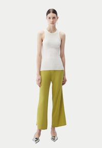 Ung kvinna som står, iklädd en vit ärmlös ribbad topp, gula ribbade vida byxor, silvriga spetsiga skor och ringörhängen.