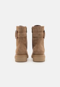 Botines de ante marrón con un acabado texturizado, que cuentan con cierre lateral de cremallera, correa ajustable con hebilla y suela de goma en color marrón claro.