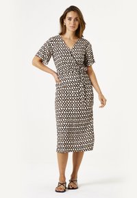 Wrap jurk in crème met een bruine ruitpatroon, korte mouwen, tailleband en een lengte tot de knie, gedragen met platte zwarte sandalen.