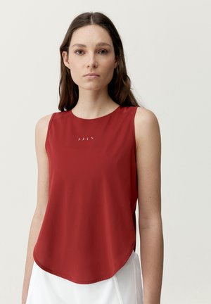 Junge Frau mit langen braunen Haaren, die ein rotes ärmelloses Top mit kleinen weißen Symbolen auf der Brust und weiße Hosen trägt, steht vor weißem Hintergrund.