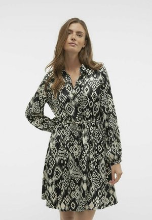 Robe à motifs noir et blanc avec des manches longues, un devant boutonné et une taille cintrée, présentant un design géométrique et une texture de tissu lisse.