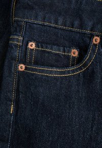 Gros plan d'une poche de jean en denim bleu foncé avec des coutures jaunes et des rivets en cuivre aux coins de la poche.