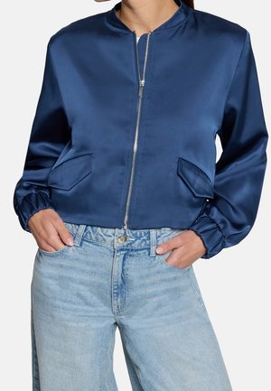 Femme portant une veste bomber bleue brillante à fermeture éclair avec poches rabatées et poignets élastiqués, associée à un jean bleu clair taille haute.