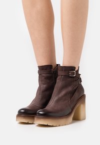 Botas de tobillo de cuero marrón con un tacón transparente grueso, cremallera lateral, detalle de hebilla y punta redondeada. Superficie texturizada y suela resistente.