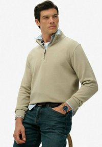 Beżowy pulower na zamek z długimi rękawami, noszony na jasnoniebieskiej koszuli z guzikami. W połączeniu z niebieskimi dżinsami i zegarkiem na nadgarstku.