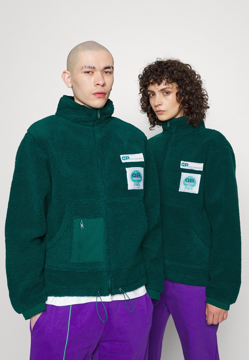 Green fleece jackets with zip pockets, high collars, and logo patches. One jacket features drawstrings at the hem, worn over purple pants.