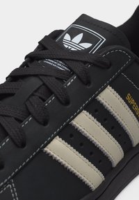 Μαύρο αθλητικό παπούτσι Adidas Superstar με λευκές ρίγες, μαύρα κορδόνια, λογότυπο στη γλώσσα και χρυσό κείμενο "Superstar" στο πλάι.