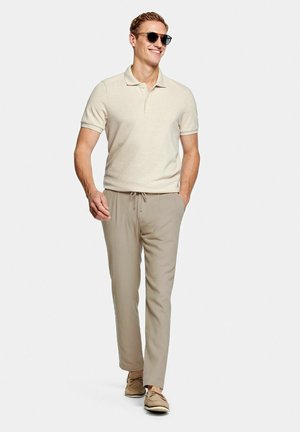 Lächelnder Mann läuft, trägt beiges Poloshirt, hellkhakifarbene Hose mit Kordelzug, Sonnenbrille und hellbraune Bootsschuhe vor einfarbigem Hintergrund.
