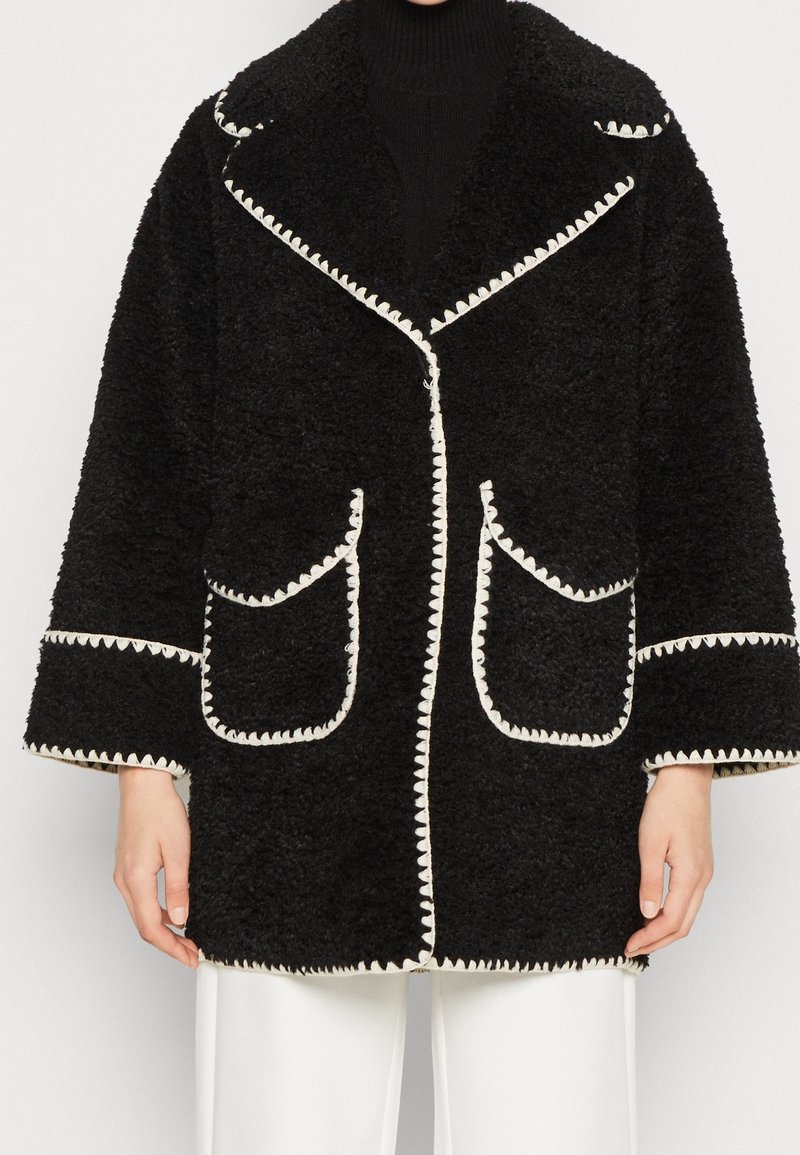 Zwarte pluizige jas met witte geschulpte rand, grote opgestikte zakken, brede kraag en oversized silhouet. Gestructureerde, warme stof.