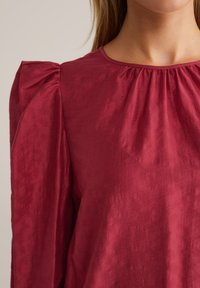 Blusa bordeaux con maniche a palloncino, scollo arricciato e una leggera texture stropicciata per un tocco di profondità.