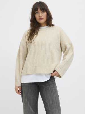 Ung kvinde med langt mørkt hår iført en beige ribstrikket sweater over en hvid skjorte og grå jeans, stående mod en ensfarvet lys baggrund.