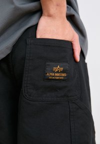 Pantalons cargo noirs en tissu texturé, avec une poche à rabat et une étiquette orange "Alpha Industries" avec un emblème.
