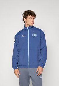 Blå vindjakke med lys blå glidelås og detaljer. Har en Adidas-logo og et grafisk motiv på venstre erme. Lettvektstoff.