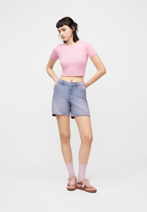 Jeune femme aux cheveux courts et foncés portant un crop top rose, un short en jean, des chaussettes roses et des baskets assorties, debout les mains dans les poches.