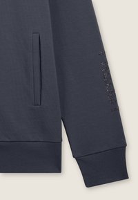 Sudadera marina con un acabado texturizado, presenta un bolsillo lateral y puños acanalados, adornada con letras de strass a lo largo de la manga.