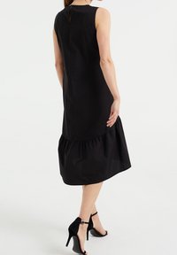 Schwarzes ärmelloses Kleid mit einem Rüschensaum, das ein dezentes Schlüsselaugen-Detail am Rücken aufweist. Die glatte Textur und der taillierte Schnitt betonen das Design.