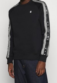 Black sweatshirt with gray accents on the sleeves. Features a crew neck, ribbed cuffs and hem, and decorative logos along the sleeves.
