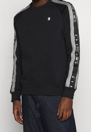 Sudadera negra con acentos grises en las mangas. Presenta un cuello redondo, puños y dobladillo acanalados, y logotipos decorativos a lo largo de las mangas.