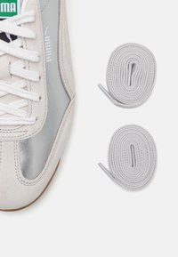 Baskets en daim gris avec accents métalliques et lacets blancs. Deux lacets ronds gris clair sont placés à côté de la chaussure.