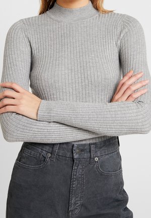 Personne portant un haut côtelé gris clair à manches longues avec un col montant et un pantalon en velours côtelé gris foncé taille haute, bras croisés.