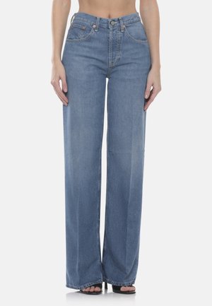 Donna che indossa jeans a vita alta, di colore azzurro chiaro, a gamba larga, abbinati a tacchi neri aperti sulle dita, in piedi davanti a uno sfondo chiaro e semplice.