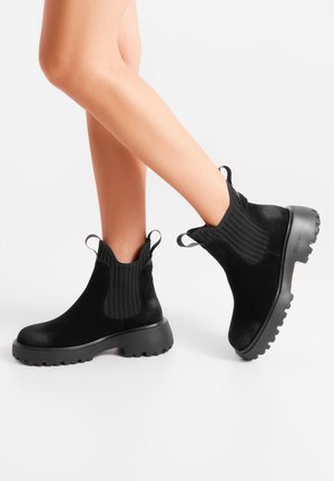 Piernas con botas Chelsea de ante negro, con suelas gruesas, paneles de elástico acanalado en el tobillo y lengüetas para tirar, sobre un fondo blanco liso.
