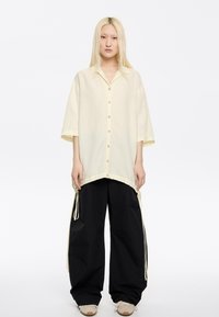 Camicia button-up giallo chiaro con vestibilità comoda, maniche corte e orlo con coulisse. Abbinata a pantaloni neri larghi e sneakers beige.