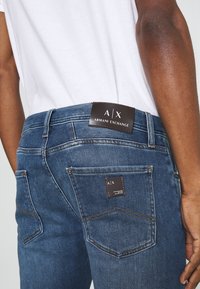 Bakifrån av en person som bär blå Armani Exchange denimjeans och en vit skjorta, som visar varumärkeslappar på midjebandet och bakfickan.