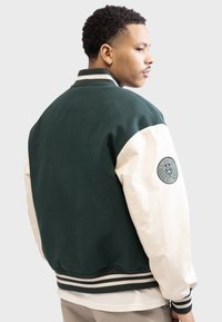Green and cream varsity jacket, wool body, synthetic sleeves, ribbed cuffs and hem, featuring a circular logo on the left sleeve.