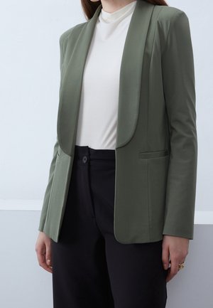 Kvinde iført en olivengrøn blazer, hvid højhalset top og sorte bukser, stående mod en lys grå baggrund.