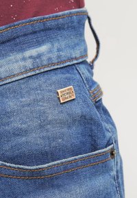 Džínové kalhoty v světle modré barvě, se kontrastním oranžovým šitím a kovovou štítkem s nápisem "PETROL Ind. Co." připevněným na zadní kapse.