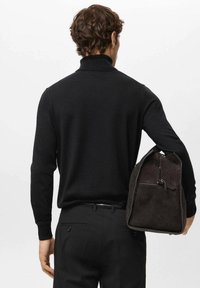 Zwarte gebreide trui met hoge kraag, gecombineerd met donkere broek. De persoon houdt een donkerbruine suède tas met ritssluiting en accentdetails vast.