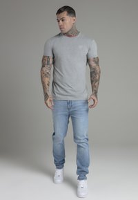 Grijze t-shirt met korte mouwen en een ronde hals, gedragen met lichtblauwe spijkerbroek en witte sneakers. Heeft zichtbare tatoeages op de armen.
