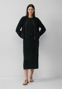 Robe plissée noire avec une taille cintrée, accompagnée d'une veste courte, zippée et ample. Les chaussures sont des sandales plates à lanières.