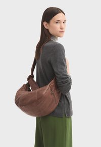 Braune Leder-Schultertasche mit einer geschwungenen Form, die eine strukturierte Oberfläche und eine Reißverschlusstasche aufweist. Das Model trägt einen grauen Pullover und grüne Hosen.
