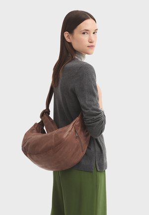 Sac à main en cuir marron avec une forme incurvée, présentant une surface texturée et une poche zippée. Le modèle porte un pull gris et un pantalon vert.