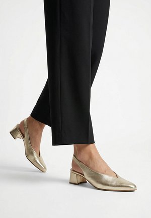 Pies con zapatos slingback dorados de tacón bloque bajo combinados con pantalones negros cortos, de pie sobre una superficie blanca.