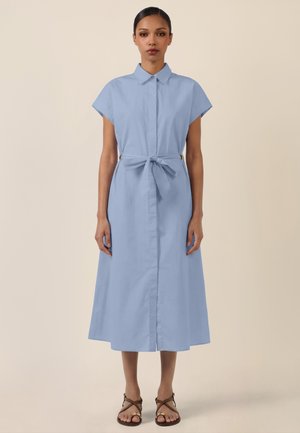 Woman wearing a light blue, short-sleeve, collared midi dress with a tied waist belt and brown flat sandals, standing against a beige background.