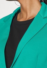 Tyrkysový blazer se strukturovaným límcem, vrstvený přes černou košili. Tkanina se zdá hladká s mírným leskem. Detailní pohled zaměřuje na linie výstřihu.