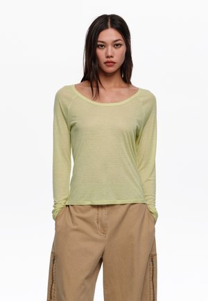 Mujer con cabello largo y oscuro viste una blusa de manga larga verde claro y pantalones cargo beige, de pie con las manos en los bolsillos contra un fondo blanco.