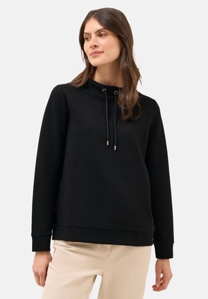Zwarte sweatshirt met een hoge hals, trekkoordsluiting, lange mouwen en ribgebreide manchetten; gecombineerd met lichtbeige broek.