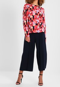 Femme portant un chemisier à manches longues aux motifs géométriques colorés, un pantalon large noir raccourci et des talons hauts noirs à bride à la cheville, sur un fond uni.