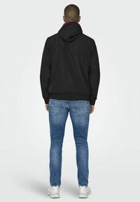 Zwarte hoodie met elastische manchetten, gecombineerd met lichtblauwe jeans. Zichtbare details op de achterkant van de jacket en denimstructuur.