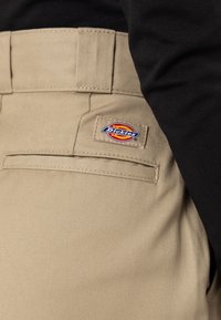 Bežové kalhoty Dickies s kapsou a logem, nošené s černým topem, ukazující texturu látky a detaily šití.
