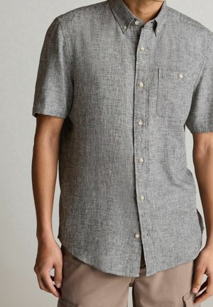 Man met een grijs gestructureerd overhemd met korte mouwen en een borstzak, en beige broek, staand tegen een neutrale achtergrond.